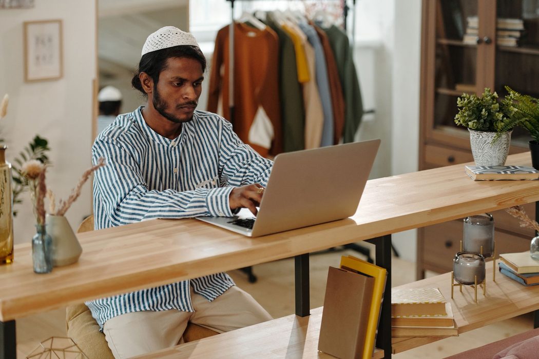 A person working at a laptop on a plain wooden desk with a notebook beside it